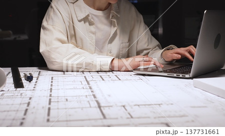 Architect or engineer working late, typing on laptop, reviewing blueprints and technical drawings for a new construction project or building plan in an office 137731661