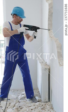 Man construction worker demolishing concrete wall with rotary hammer, wearing protective safety gear, generating dust and debris during home renovation work Man construction worker demolishing concrete wall with rotary hammer, wearing protective safety gear, generating dust and debris during home renovation work 137731686
