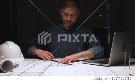 Architect working late into the night, focusing on detailed blueprints and construction plans using a ruler, illuminated by a focused spotlight on the desk Architect working late into the night, focusing on detailed blueprints and construction plans using a ruler, illuminated by a focused spotlight on the desk 137731739