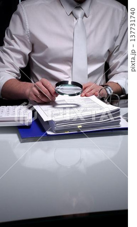 Professional businessman analyzing financial reports and charts with a magnifying glass while working at a desk. Audit and taxes theme in business Professional businessman analyzing financial reports and charts with a magnifying glass while working at a desk. Audit and taxes theme in business 137731740