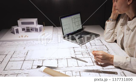 Female architect facing burnout after long late nights, rubbing tired eyes while holding glasses amid blueprints, laptop and a modern building model on her cluttered desk 137731800