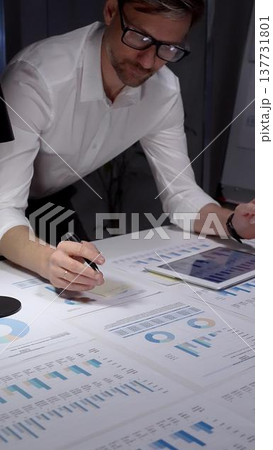 Business professional studies financial reports and market data at a cluttered desk, surrounded by charts, graphs and a laptop, focused on planning, strategy and results 137731801