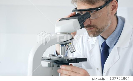 Adult male scientific researcher analyzing sample through microscope, wearing protective gear in high tech laboratory environment. Healthcare, medicine and science concept 137731904
