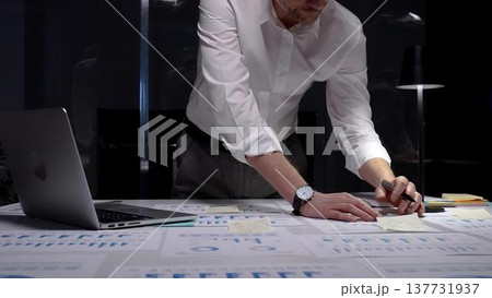 Professional businessman standing at a brightly lit desk, focused on writing notes and reviewing financial reports, charts and data late into the night to meet deadlines Professional businessman standing at a brightly lit desk, focused on writing notes and reviewing financial reports, charts and data late into the night to meet deadlines 137731937
