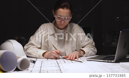 Young woman architect working late at laptop, studying blueprints and design plans on a desk with hard hat and rolled drawings, focused on construction planning and deadlines 137731985