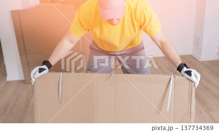 A worker unpacks furniture from cardboard boxes. Man lifts large cardboard box with effort. Hands grip sides, wearing white gloves. Bright yellow shirt contrasts with soft lighting. Gentle smile 137734369