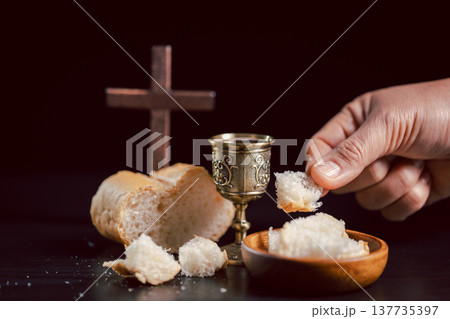 Communion scene with hand offering bread beside ornate chalice and wooden cross on dark background, symbolizing Christian Eucharist, worship, faith, sacrifice and remembrance with copy space. 137735397