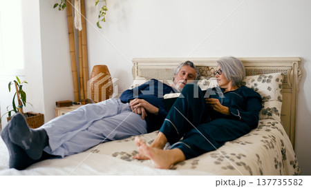 Senior couple relaxing in bed reading book together Senior couple relaxing in bed reading book together 137735582