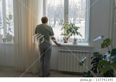 Elderly woman standing by a window, looking out at the snow-covered trees during a cold winter day, feeling solitude Elderly woman standing by a window, looking out at the snow-covered trees during a cold winter day, feeling solitude 137741345