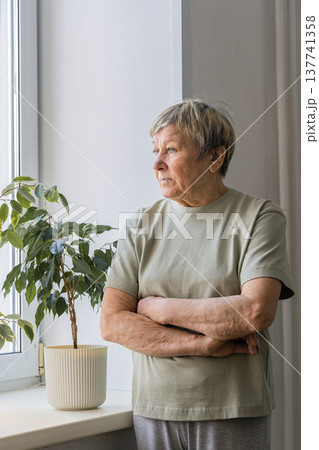 Senior woman standing by window with arms crossed, thoughtful and pensive in quiet home light, reflecting on solitude and memory Senior woman standing by window with arms crossed, thoughtful and pensive in quiet home light, reflecting on solitude and memory 137741358