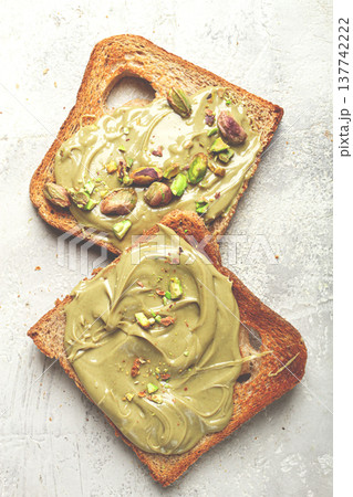 Two slices of toasted bread, topped with pistachio paste and sprinkled with pistachios, spread on a light surface Two slices of toasted bread, topped with pistachio paste and sprinkled with pistachios, spread on a light surface 137742222