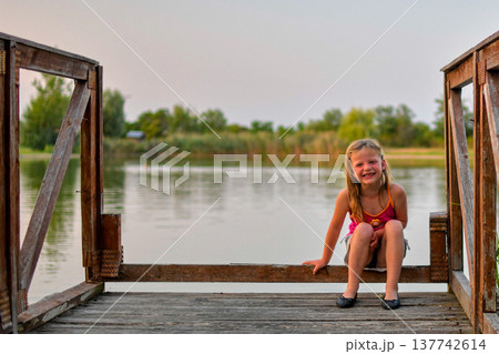 Young girl smiling on a wooden dock by a calm lake during golden hour Young girl smiling on a wooden dock by a calm lake during golden hour 137742614