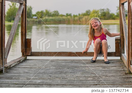 Joyful young girl laughing on a wooden pier by the tranquil lake during golden hour. A happy child with blonde hair sits on a weathered wooden dock, her face lit up with laughter, with a calm lake and 137742734