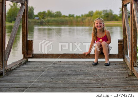 Joyful young girl laughing on a wooden dock by the tranquil lake at sunset. A happy child with blonde hair sits on a weathered wooden pier, her face lit up with laughter as she enjoys a peaceful Joyful young girl laughing on a wooden dock by the tranquil lake at sunset. A happy child with blonde hair sits on a weathered wooden pier, her face lit up with laughter as she enjoys a peaceful 137742735