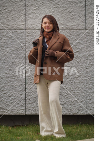 A woman dressed for autumn looks into the distance, her brown coat and scarf highlighting comfort and maturity. The photo captures warmth and classic seasonal fashion. 137742849
