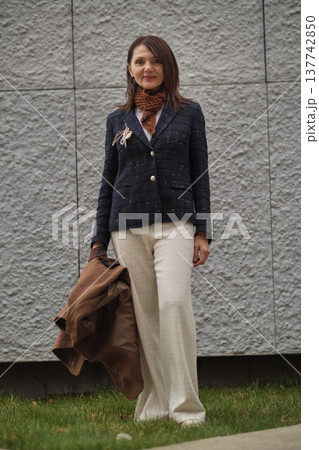 A woman holds a brown coat and looks away with a gentle smile. The image highlights mature confidence, autumn mood, and effortless elegance in everyday fashion. A woman holds a brown coat and looks away with a gentle smile. The image highlights mature confidence, autumn mood, and effortless elegance in everyday fashion. 137742850