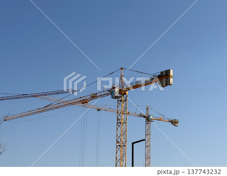 A tower crane stretches across the sky above an active construction area. Infrastructure, progress, urban growth, engineering ambition, industrial scale, and the clean geometry of structural A tower crane stretches across the sky above an active construction area. Infrastructure, progress, urban growth, engineering ambition, industrial scale, and the clean geometry of structural 137743232