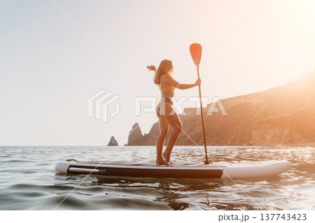Woman Paddleboarding Ocean Coastline - A woman paddleboards in the ocean with a scenic coastline in the background. Woman Paddleboarding Ocean Coastline - A woman paddleboards in the ocean with a scenic coastline in the background. 137743423
