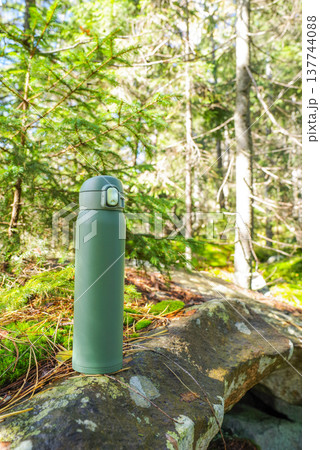 Green thermos on the stone. Thermal bottle on the background of the forest. Sunny day in the forest. Green thermos on the stone. Thermal bottle on the background of the forest. Sunny day in the forest. 137744088