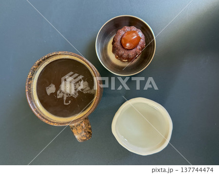 Coffee cup with dessert canele and glass of water served on dark table in cafe setting. Cafe food presentation, coffee break concept, restaurant menu content, lifestyle dining Coffee cup with dessert canele and glass of water served on dark table in cafe setting. Cafe food presentation, coffee break concept, restaurant menu content, lifestyle dining 137744474