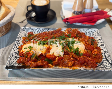 Shakshuka with eggs and herbs served beside coffee and bread. Hearty breakfast, spice, cafe comfort, shared dining, and the rich warmth of a leisurely morning meal. 137744677