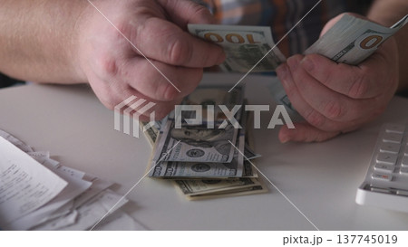 A person sits at a table counting bills. Receipts are scattered on the surface, and the light indicates it is daytime. The focus is on the act of counting cash. 137745019