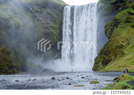 Huge waterfall surrounded by mossy rocky cliff and people walking towards it, Iceland 137745052