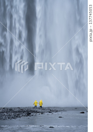 Vertical shot of couple in yellow raincoats walking in front of huge waterfall, Iceland 137745053