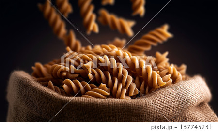 Close-up of whole wheat fusilli pasta falling into a rustic burlap sack on dark background 137745231