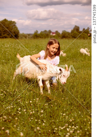 Girl cradling a baby goat in her arms, surrounded by a lush green field, basking in the warmth of a sunny day 137745399