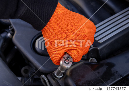 A man in an orange glove focuses intently on repairing a car engine, showcasing skill and dedication in a garage setting 137745877