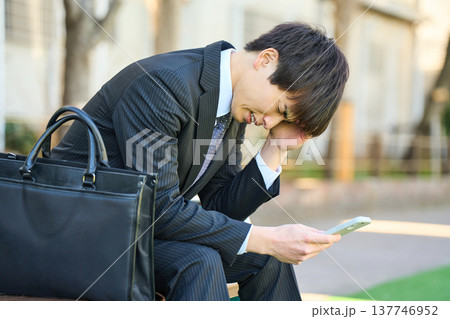 園のベンチでスマホを見て落ち込む若いビジネスマン 上半身 137746952