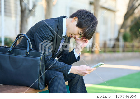 園のベンチでスマホを見て落ち込む若いビジネスマン 上半身 園のベンチでスマホを見て落ち込む若いビジネスマン 上半身 137746954