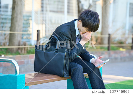 園のベンチでスマホを見て落ち込む若いビジネスマン 上半身 園のベンチでスマホを見て落ち込む若いビジネスマン 上半身 137746965