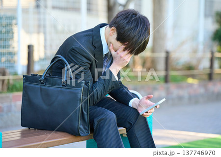 園のベンチでスマホを見て落ち込む若いビジネスマン 上半身 園のベンチでスマホを見て落ち込む若いビジネスマン 上半身 137746970