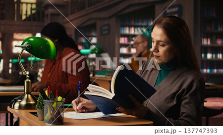 Determined woman writing thesis citations from a specialty book at the library, mature learner working toward a higher education goal. Academic materials in a quiet campus study area. Determined woman writing thesis citations from a specialty book at the library, mature learner working toward a higher education goal. Academic materials in a quiet campus study area. 137747393