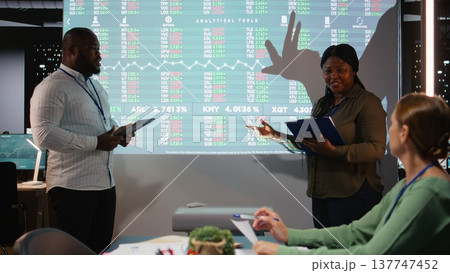 African american associates presenting their pitch to shareholders in a late night meeting, discussing about the financial situation and working for success. Evaluate supply and demand. Camera A. 137747452