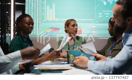 Close up of diverse staff hand over data projections papers in a meeting after hours for multinational company operations. Risk assessment and visual data to support forecasting. Camera A. Close up of diverse staff hand over data projections papers in a meeting after hours for multinational company operations. Risk assessment and visual data to support forecasting. Camera A. 137747487