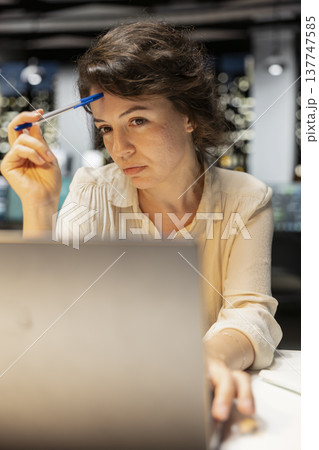Businesswoman contemplating on complex problem in dark office late at night, strategizing and typing options on laptop. Seeking a logical solution through methodical concentration. 137747585