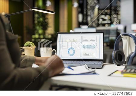 African american skilled analyst reviewing monthly reports for a new growth strategy, using infographics on laptop to plan resources allocation and align objectives with financial development. 137747602