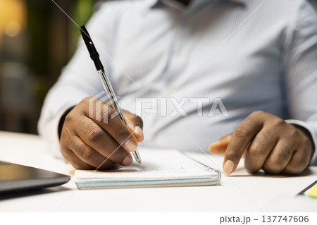 Close up of black male employee documenting essential ideas on textbook, scheduling tasks and doing research at midnight. Drafting handwritten notes for presentation that reflects objectives. Close up of black male employee documenting essential ideas on textbook, scheduling tasks and doing research at midnight. Drafting handwritten notes for presentation that reflects objectives. 137747606