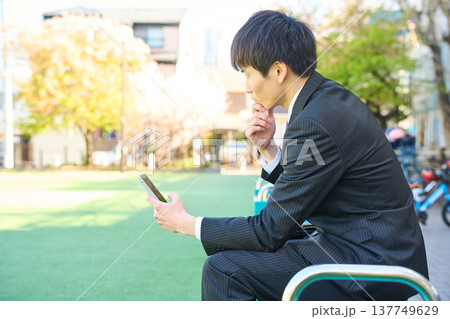 公園のベンチに座り真剣な表情でスマホを見る新人ビジネスマン 上半身 137749629