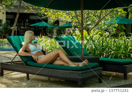 Woman lounges beside the pool in a lush garden setting Woman lounges beside the pool in a lush garden setting 137751281