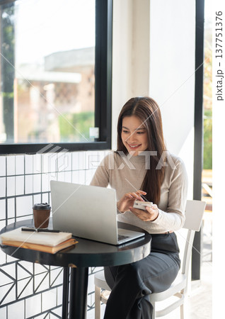 Smiling asian woman holding phone looking at laptop aside books and coffee as sits at table in cafe. 137751376