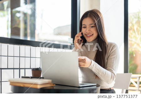 Smiling asian woman talking on phone pointing at laptop aside books and coffee sit at table in cafe. 137751377