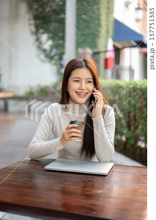 Smiling asian woman talking on phone holding coffee over laptop sitting at wooden table outside cafe Smiling asian woman talking on phone holding coffee over laptop sitting at wooden table outside cafe 137751385