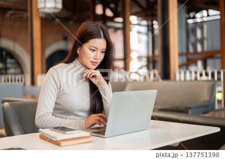Pretty asian woman putting hand under chin looking at laptop aside phone and book on table in cafe. 137751398