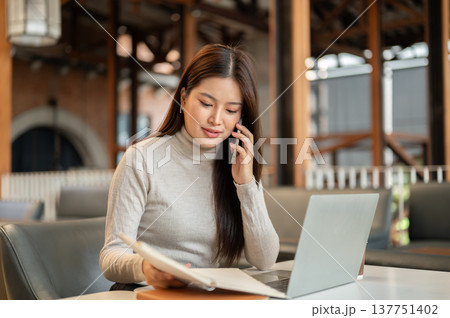 Pretty asian woman talking on phone as holding notebook aside laptop while sitting at table in cafe. 137751402