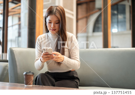 Pretty asian woman looking and using smartphone aside coffee on table while sitting on sofa in cafe. Pretty asian woman looking and using smartphone aside coffee on table while sitting on sofa in cafe. 137751455