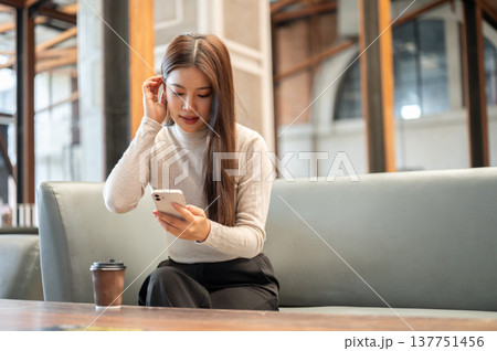 Pretty asian woman looking at phone and tucking hair aside coffee on table sitting on sofa in cafe. Pretty asian woman looking at phone and tucking hair aside coffee on table sitting on sofa in cafe. 137751456